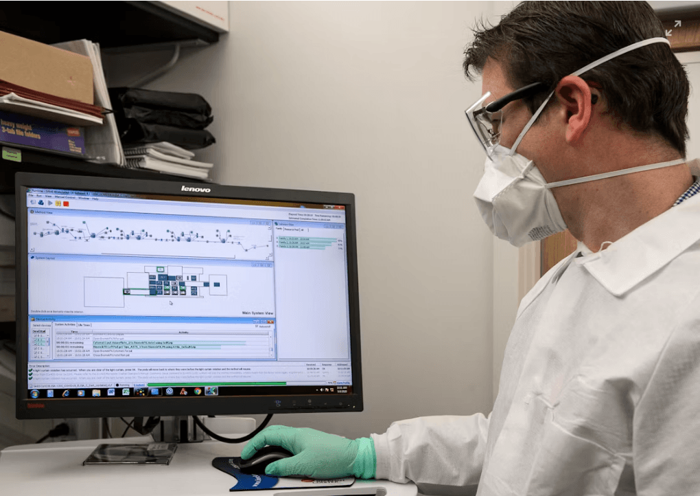 A laboratory technician in a lab coat and mask reviewing results on a computer.